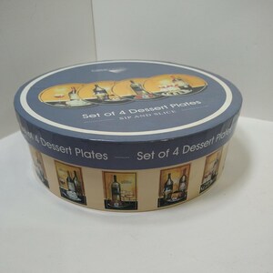 May include: A round box set of 4 dessert plates. The box is blue and tan with images of wine bottles and glasses. The text "Set of 4 Dessert Plates" and "SIP AND SLICE" is printed on the box.