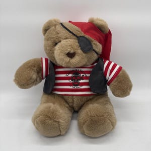 May include: A light brown plush teddy bear dressed as a pirate. The bear wears a red headscarf, a black eye patch, and a red and white striped shirt with a black skull graphic and the text Treasure Island at the Mirage, plus a black vest.