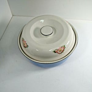 May include: A round, light blue enamel bowl with a matching cream-colored lid. The lid features a central handle and a delicate floral pattern with pink flowers and green leaves. The bowl has a dark rim and a smooth, glossy finish throughout.