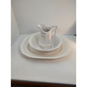 May include: A white ceramic wash basin set. The set includes a pitcher, a bowl, and a rectangular tray. The pieces have a simple, classic design, with subtle detailing. The set is likely for decorative or functional use.