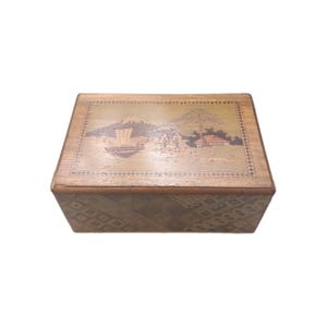 May include: Wooden box with a landscape scene on the lid. The box features a detailed illustration of a mountain, boat, and buildings. The sides of the box have a geometric pattern. The box is rectangular.
