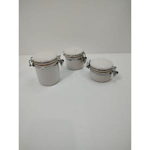 May include: Three white ceramic canisters with rounded lids and metal clasps. The canisters are different sizes, with a light beige band around the middle. The lids are white and have a slight gloss.