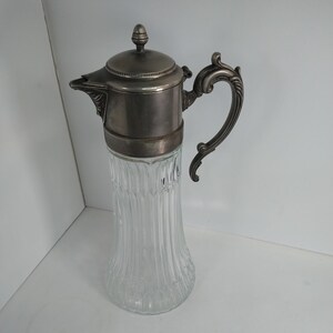 May include: A vintage clear glass pitcher with a silver-colored metal lid, handle, and spout. The glass body has a vertical ribbed design. The handle is ornate. The pitcher is likely used for serving beverages.