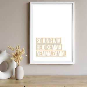 Può includere: Una stampa incorniciata bianca con il testo "SO JUNG WIA HEID KEMMA NEMMA ZAMM." in beige su sfondo bianco. La stampa è appesa a una parete beige chiaro sopra un tavolo di legno con due vasi e fiori secchi.