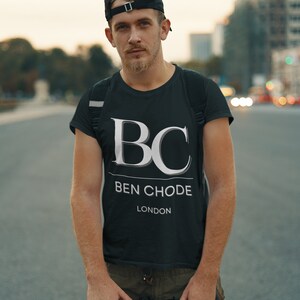 Puede incluir: Un hombre con una camiseta negra con un logotipo blanco "BC BEN CHODE LONDON", una gorra de béisbol negra y pantalones cargo color caqui. Está sujetando el manillar de una bicicleta negra.