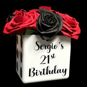 May include: A white cube-shaped vase with black polka dots and the text "Sergio's 21st Birthday" in black lettering. The vase is filled with red and black roses.