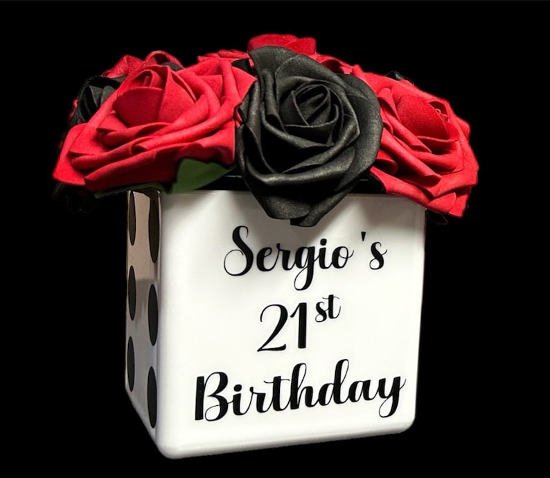 May include: A white cube-shaped vase with black polka dots and the text "Sergio's 21st Birthday" in black lettering. The vase is filled with red and black roses.