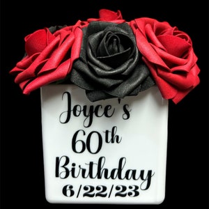 May include: A white ceramic vase with black lettering that reads "Joyce's 60th Birthday 6/22/23". The vase is filled with red and black artificial roses.