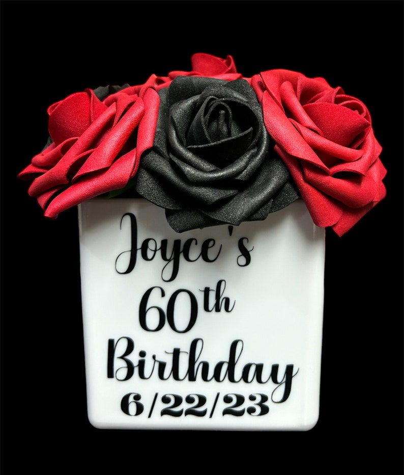 May include: A white ceramic vase with black lettering that reads "Joyce's 60th Birthday 6/22/23". The vase is filled with red and black artificial roses.