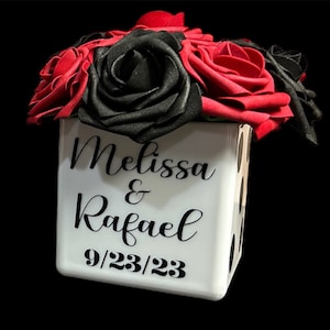 May include: A white cube vase with black polka dots, decorated with the names "Melissa & Rafael" and the date "9/23/23". The vase is filled with a bouquet of red and black roses.