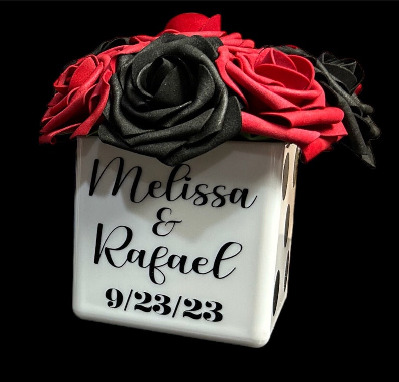 May include: A white cube vase with black polka dots, decorated with the names "Melissa & Rafael" and the date "9/23/23". The vase is filled with a bouquet of red and black roses.