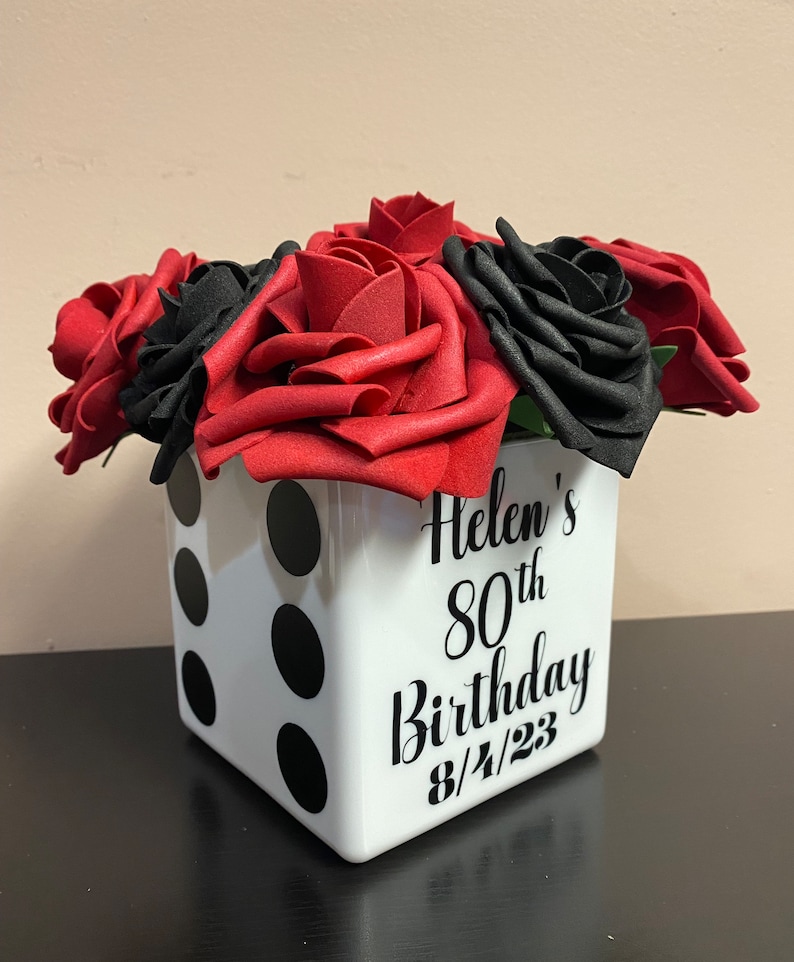 May include: A white cube-shaped vase with black dots, resembling a dice, decorated with the text "Helen's 80th Birthday 8/4/23". The vase is filled with red and black foam roses.