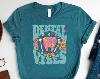 May include: A teal blue t-shirt featuring the text "Dental Vibes" in a retro font. The text is surrounded by flowers and a pink tooth with dental tools. The shirt is laid flat on a white surface with a pair of glasses, a wallet, a camera, and a plant in a pot.