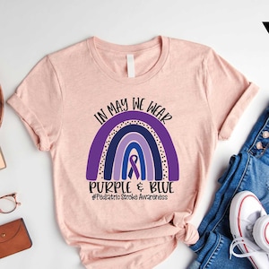 May include: A heather peach t-shirt with a rainbow design and the text "In May We Wear Purple & Blue #Pediatric Stroke Awareness".