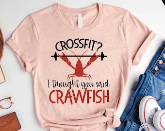 May include: A pink t-shirt with a black and white graphic of a barbell and a red crawfish. The text on the shirt reads "Crossfit? I thought you said Crawfish".