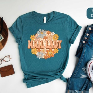 May include: Teal t-shirt with the text "MAIL LADY" in an orange retro font, surrounded by a floral design. The shirt is paired with blue jeans and white sneakers. The image also includes a wooden tray, a camera, and glasses.