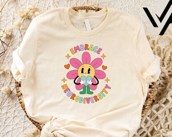 May include: A cream colored t-shirt with a colorful graphic of a smiling flower holding a heart. The text "Embrace Neurodiversity" is written in a rainbow font around the flower.