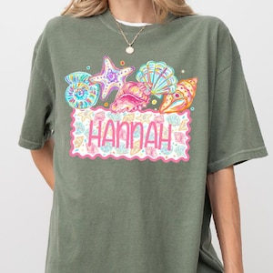 May include: Moss green t-shirt featuring a colorful graphic of seashells, a starfish, and the name "Hannah" in a pink scalloped frame. The shirt is a relaxed fit and has short sleeves.