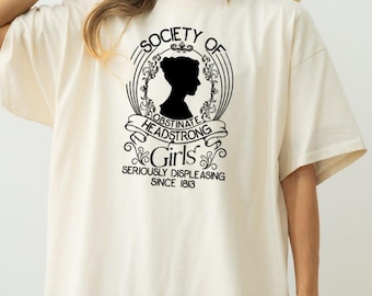 May include: An ivory short-sleeved t-shirt featuring a black graphic print on the chest. The design includes a silhouette of a person inside an oval frame with the text: SOCIETY OF OBSTINATE HEADSTRONG Girls SERIOUSLY DISPLEASING SINCE 1813.