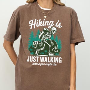 May include: Espresso-colored t-shirt with the text "Hiking is Just Walking Where You Might Die." The graphic features a skeleton hiking with a bear and a backpack. The shirt is a comfortable, casual style.