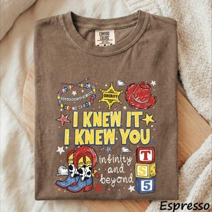 A t-shirt with a cartoon-style design featuring a cowboy hat, a sheriff's badge, and various other cartoon-style elements. The text on the t-shirt reads "I KNEW IT I KNEW YOU in infinity and beyond" and includes a reference to the character Toy Story.