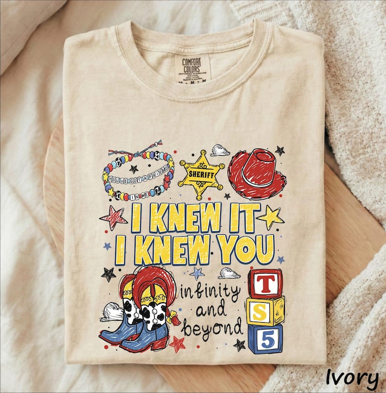 A beige t-shirt with a colorful graphic design featuring various cartoon-style icons and text, including a cowboy hat, star, and the phrase "I KNEW IT" in yellow.