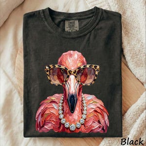May include: A charcoal black t-shirt featuring a painterly illustration of a pink flamingo wearing leopard print sunglasses and a silver-toned rhinestone necklace. The graphic has visible brushstroke textures and a central placement on the chest.