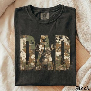 May include: Black t-shirt with the word "DAD" in camouflage print, featuring a deer graphic. The shirt has a crew neck and short sleeves. The text "Black" is at the bottom right corner.