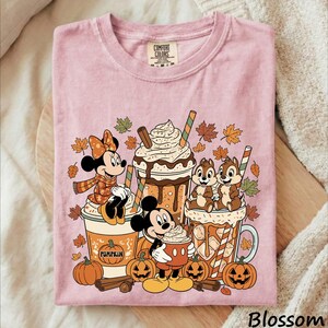May include: A light pink t-shirt featuring a graphic print of Minnie Mouse, Mickey Mouse, and Chip 'n Dale with autumn leaves, pumpkins, and three large beverage cups. The text COMFORT COLORS is printed on the inside neck label of the garment.