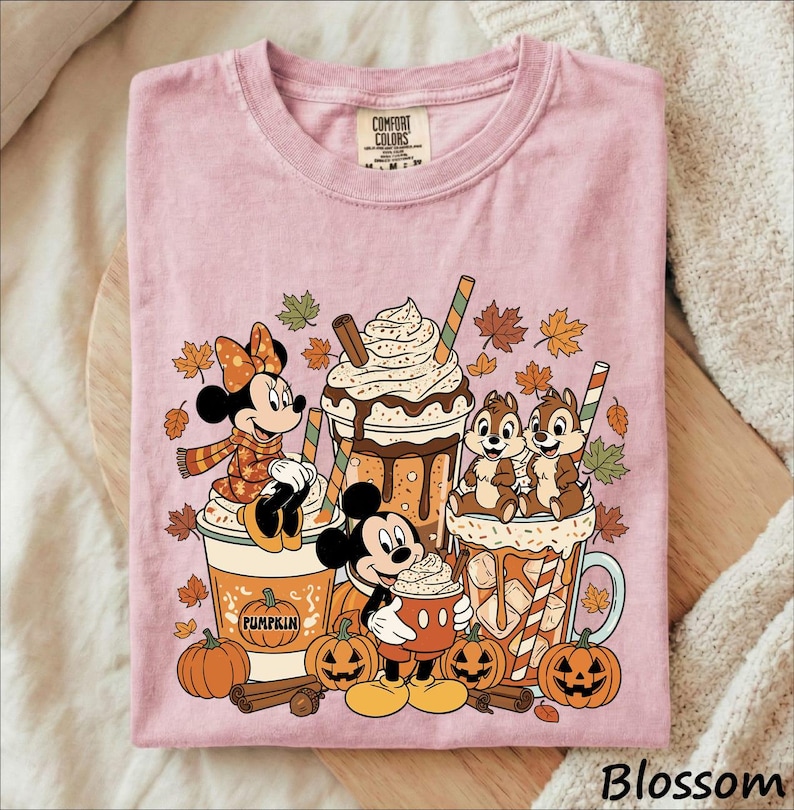 May include: A light pink t-shirt featuring a graphic print of Minnie Mouse, Mickey Mouse, and Chip 'n Dale with autumn leaves, pumpkins, and three large beverage cups. The text COMFORT COLORS is printed on the inside neck label of the garment.
