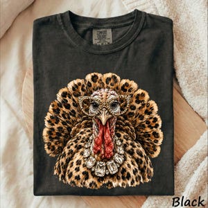 May include: A charcoal black crew neck t-shirt featuring a graphic of a turkey with leopard print feathers, wearing leopard print glasses and a large crystal-style necklace. The shirt has a Comfort Colors label printed on the inside neck area.