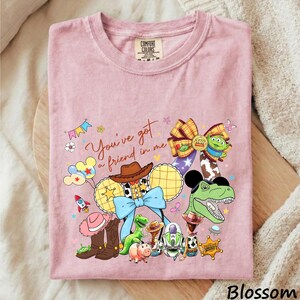 A pink t-shirt with a cartoon-style illustration of various characters from the Toy Story franchise, including Woody, Buzz Lightyear, and other beloved characters.