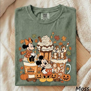 May include: A moss green t-shirt featuring a graphic print of Minnie Mouse, Mickey Mouse, Chip and Dale with autumn-themed beverages, pumpkins, cinnamon sticks, and falling leaves. The inner neck label displays the text Comfort Colors.