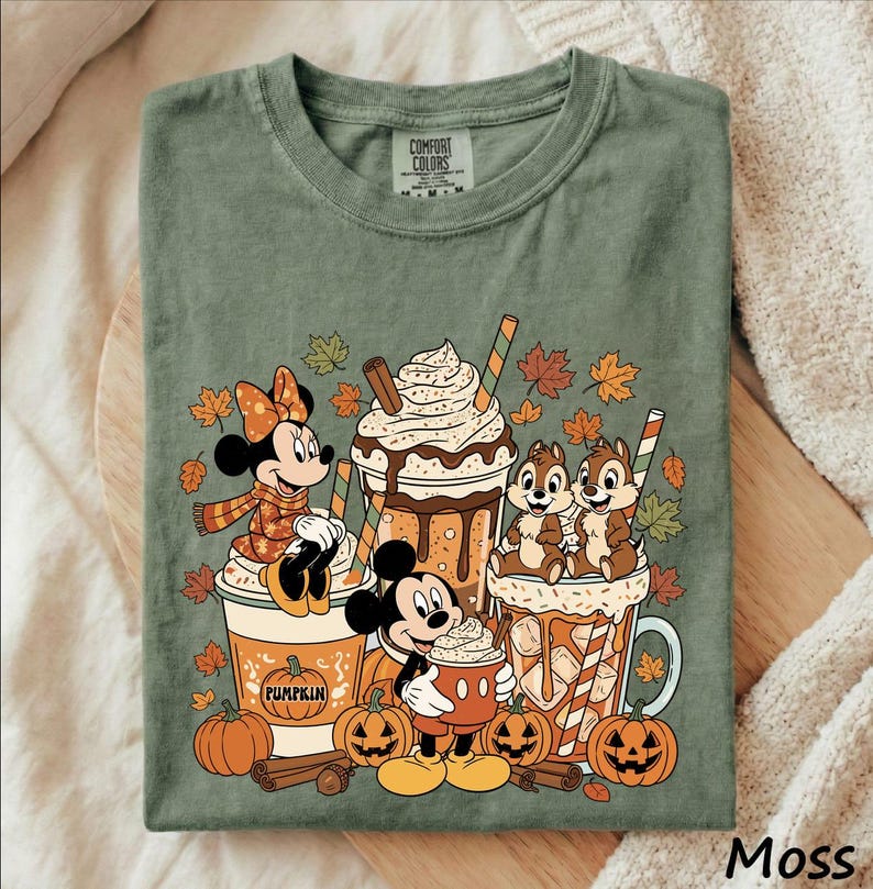 May include: A moss green t-shirt featuring a graphic print of Minnie Mouse, Mickey Mouse, Chip and Dale with autumn-themed beverages, pumpkins, cinnamon sticks, and falling leaves. The inner neck label displays the text Comfort Colors.
