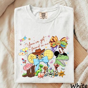 A white t-shirt with a colorful graphic design featuring various characters from the Toy Story franchise, including Woody, Buzz Lightyear, and other iconic figures.