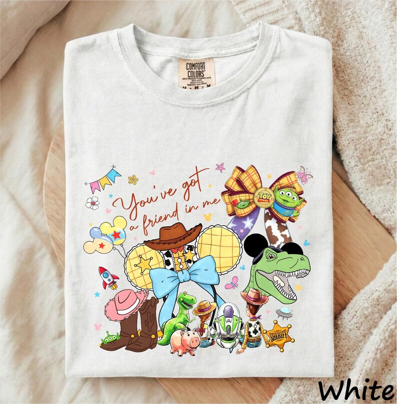 A white t-shirt with a colorful graphic design featuring various characters from the Toy Story franchise, including Woody, Buzz Lightyear, and other iconic figures.