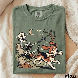 May include: A moss green crew neck t-shirt featuring a graphic print of a skeleton emerging from a grave and a beagle dog running with a bone in its mouth. The design includes a crescent moon, bats, and a bare tree. The shirt has a Comfort Colors label.
