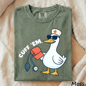 May include: Moss green t-shirt with a cartoon goose wearing sunglasses and a nurse's hat, holding a blood pressure cuff. The text "CUFF 'EM" is printed on the shirt. The shirt is folded on a wooden surface.