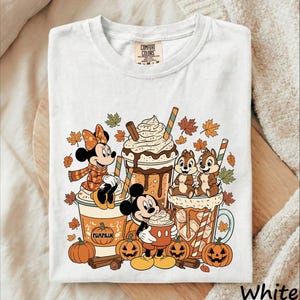 May include: A white t-shirt featuring a graphic print of Minnie Mouse, Mickey Mouse, and Chip 'n Dale with autumn-themed drinks. The design includes falling leaves, pumpkins, and cinnamon sticks. The shirt has a Comfort Colors brand neck label.