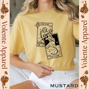May include: A mustard yellow t-shirt with a black and white illustration of two tarot cards, The Star and The Moon. The text on the cards reads "The Star" and "The Moon".