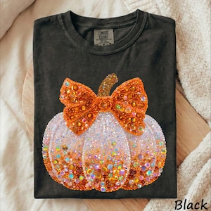 May include: A black short-sleeved t-shirt featuring a large graphic of a pumpkin adorned with orange and clear rhinestones. The pumpkin is topped with a prominent bow made of matching orange crystals. The shirt has a Comfort Colors brand label.