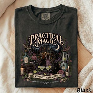 May include: A charcoal gray t-shirt featuring a graphic print with the text "PRACTICAL MAGIC" and "MIDNIGHT MARGARITAS THERE'S A LITTLE WITCH IN ALL OF US." The design includes a house, two figures, a black cat, a blender, and potion bottles.