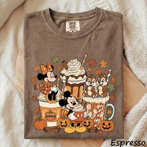 May include: A brown t-shirt featuring a graphic of Minnie Mouse, Mickey Mouse, and Chip 'n Dale with pumpkin spice lattes and jack-o'-lanterns. The design includes autumn leaves, whipped cream, and the text Comfort Colors on the inner neck label.