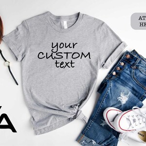 May include: A gray t-shirt with the text "your custom text" printed on it. The shirt is laid out on a white surface with a pair of jeans, a watch, and a pair of glasses. The t-shirt is labeled "Athletic Heather".