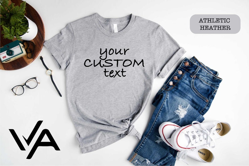 May include: A gray t-shirt with the text "your custom text" printed on it. The shirt is laid out on a white surface with a pair of jeans, a watch, and a pair of glasses. The t-shirt is labeled "Athletic Heather".