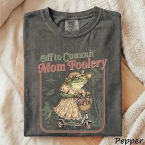 May include: Pepper-colored t-shirt with the text "Off to Commit Mom Foolery." The graphic features a frog wearing a dress and hat, riding a scooter with a basket of flowers and a teddy bear.