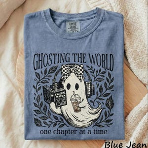 May include: A blue crew neck t-shirt featuring a graphic of a ghost wearing a checkered bow, reading a book, and holding an iced coffee. The text reads GHOSTING THE WORLD and one chapter at a time. The shirt has a Comfort Colors brand label.