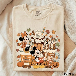 May include: A beige t-shirt featuring a graphic print of Minnie Mouse, Mickey Mouse, and Chip 'n Dale with autumn-themed drinks, pumpkins, cinnamon sticks, and falling leaves. The design includes a pumpkin illustration with the text PUMPKIN.