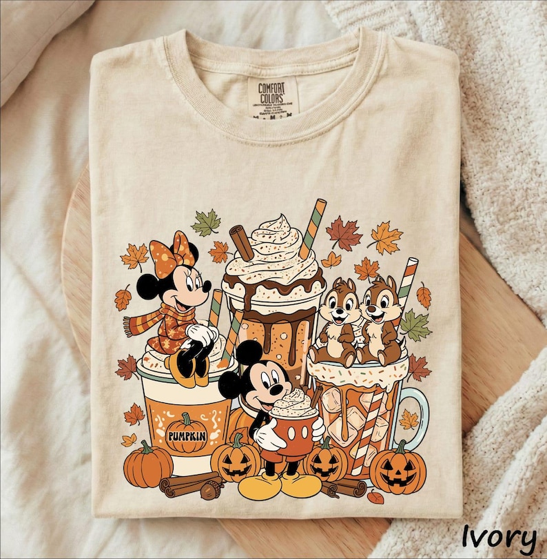 May include: A beige t-shirt featuring a graphic print of Minnie Mouse, Mickey Mouse, and Chip 'n Dale with autumn-themed drinks, pumpkins, cinnamon sticks, and falling leaves. The design includes a pumpkin illustration with the text PUMPKIN.