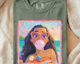 May include: An olive green t-shirt featuring a rectangular graphic print of a person with long dark hair wearing orange heart-shaped sunglasses and blowing a pink bubble. The print includes a beach background and a pink leopard print border.